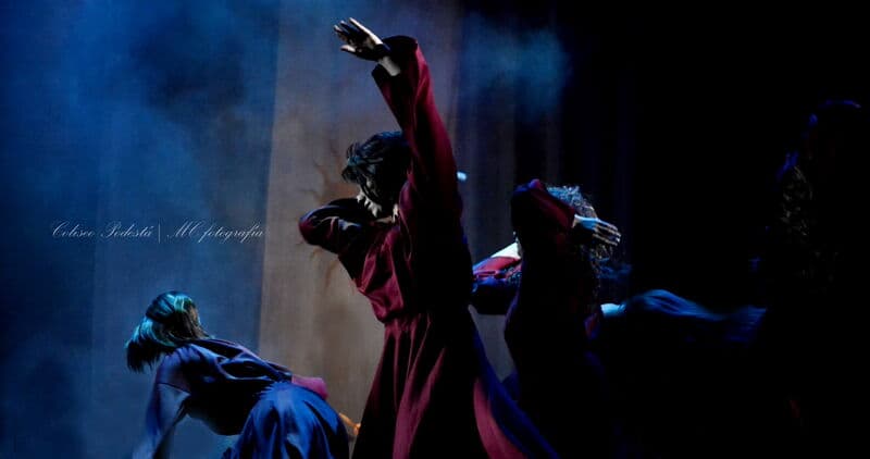 Three dancers in flowing crimson robes move through blue stage smoke, one with an arm raised high.