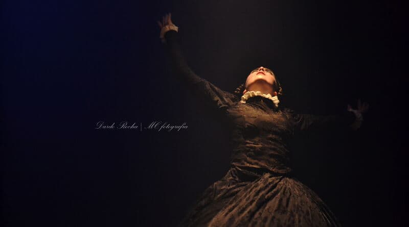 A dancer in a dark period costume with a ruffled collar tilts her head back, one arm raised against black.