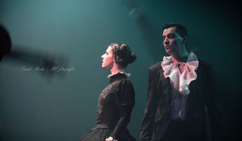 A woman in a lace collar and a man in a ruffled cravat stand in profile under teal stage light.
