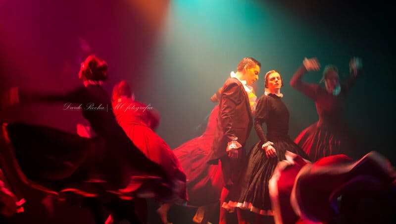 A couple in Victorian-style costume stands sharp in focus amid a blur of red-skirted dancers spinning around them.