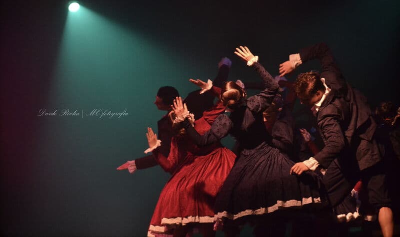A cluster of dancers in dark period costume raise their hands together under a single spotlight.