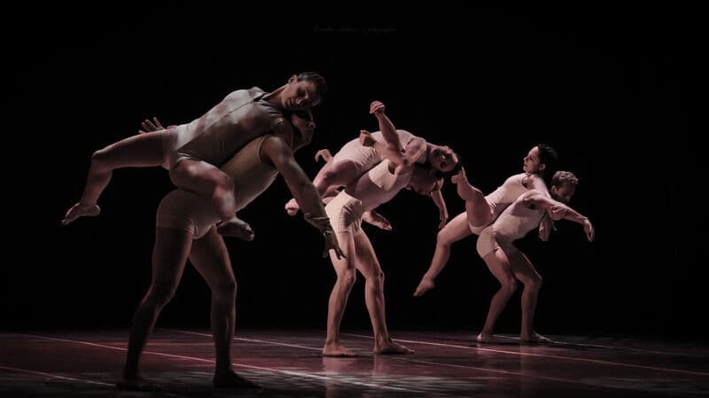 Three pairs of contemporary dancers lift and carry each other in matching nude-toned costumes against black.