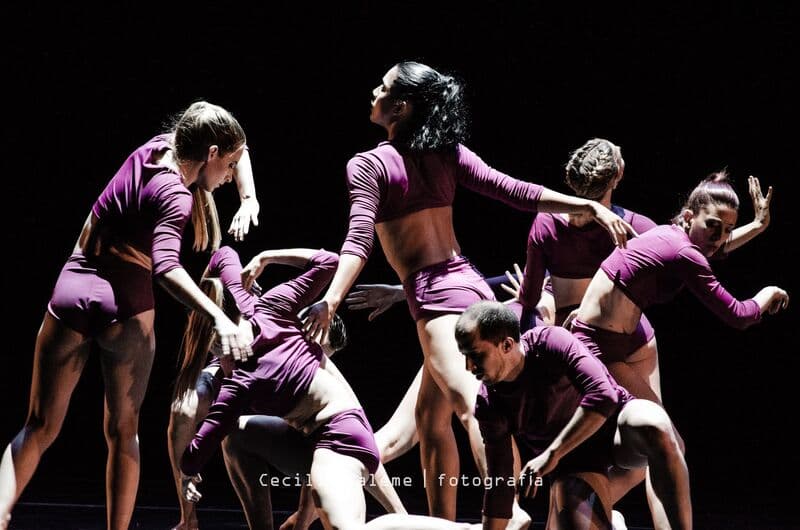 A tight cluster of dancers in magenta bodysuits reach and twist together under warm stage light.