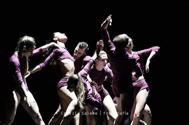 Dancers in magenta bodysuits arch and reach in a tight group, one woman's lit face leaning back.