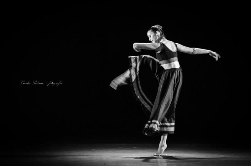 A solo dancer in a dark skirt turns on pointe under a single spotlight against black, in black and white.