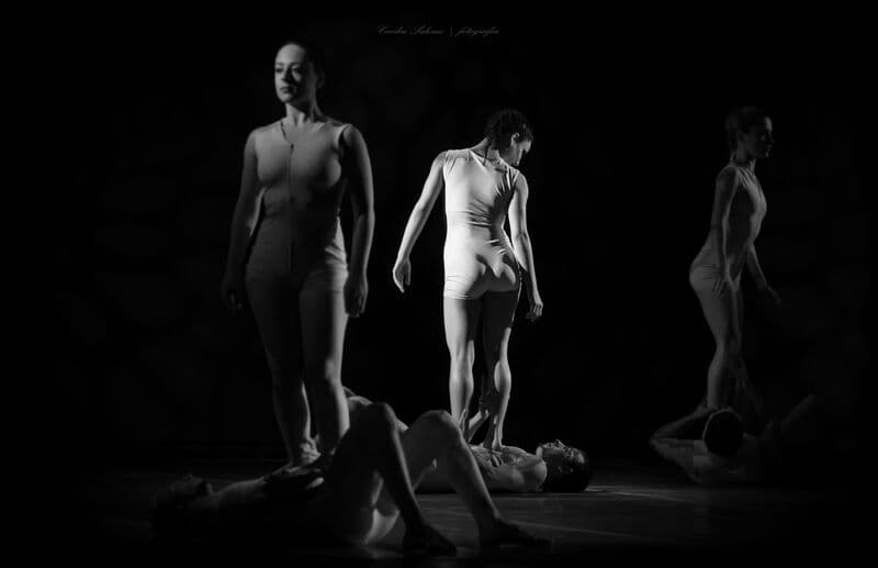 Dancers stand and balance over reclining partners on a dark stage, in black and white.