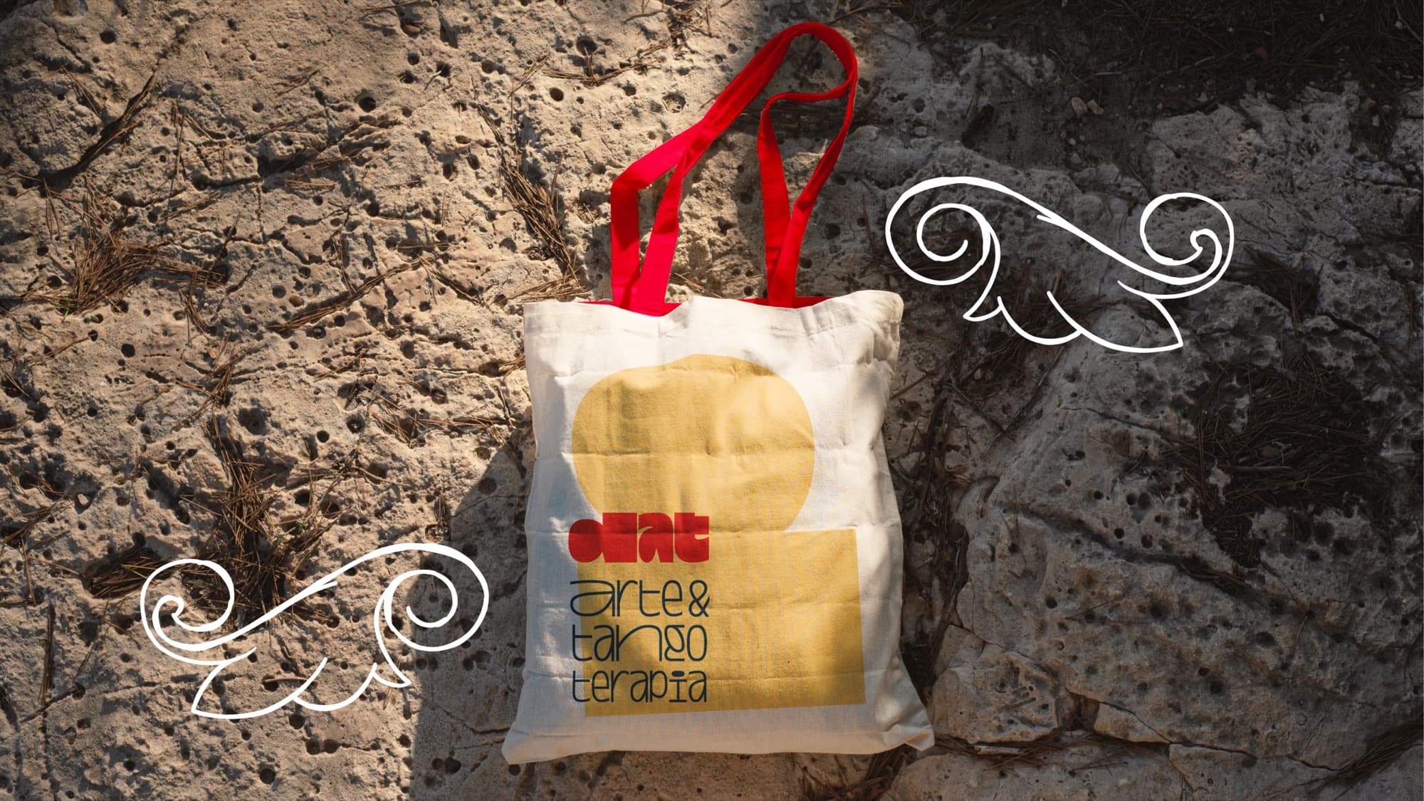 A cream tote bag with red handles and the DAT logo over a yellow shape, lying on stone, with hand-drawn flourishes around it.